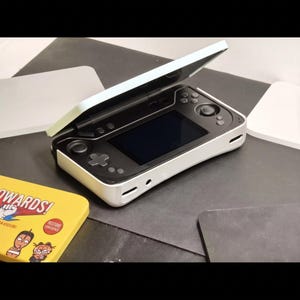 May include: A white and black handheld gaming console with a clamshell design, featuring a central screen, a directional pad, dual analog sticks, and various action buttons. The device is partially open, revealing the internal control layout.