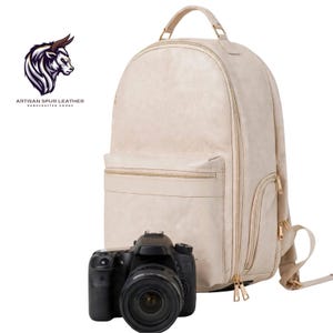 May include: A cream-colored leather backpack with gold-tone zippers and a top handle, positioned behind a black DSLR camera. The logo for Artisan Spur Leather Handcrafted Goods featuring a bull head illustration is visible in the upper left corner.