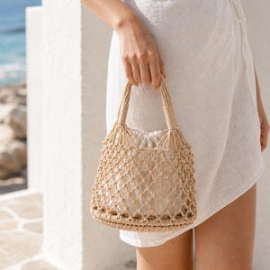 Op de afbeelding: Een beige macramé handtas met een geknoopt netontwerp en een effen witte stoffen binnenzak met trekkoord. De tas heeft twee geïntegreerde touwhengsels en een ronde bodem, vervaardigd van natuurlijk koord in een gestructureerde vorm.