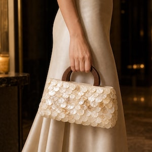 May include: A rectangular handbag featuring a surface covered in overlapping, circular, iridescent shell-like discs in shades of cream and beige. The bag is equipped with a dark brown, semi-circular wooden handle attached to the top frame.