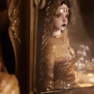 Puede incluir: Una muñeca articulada con cabello oscuro y labios oscuros, vestida con un vestido metálico dorado y un tocado a juego con detalles dorados colgantes. La muñeca está colocada dentro de una estructura con marco dorado, reflejando una luz cálida.
