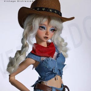 May include: A ball-jointed doll with white braided hair wearing a brown cowboy hat with a star-studded black band, a red neckerchief, and a distressed blue denim crop top. The text 'Vintage Distressed' is visible in the upper left corner.
