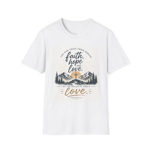Puede incluir: Una camiseta blanca de manga corta con un estampado gráfico de montañas, una cruz y un sol. El texto dice: And now these three remain: faith, hope and love. But the greatest of these is love. 1 Corinthians 13:13 en negro y dorado.