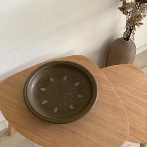 Peut inclure: Un bol en céramique vert olive foncé, de forme ronde, présentant un motif central en étoile gravé avec des accents bleu clair. Le bol possède une finition mate lisse et un bassin large et peu profond, révélant un design géométrique détaillé.