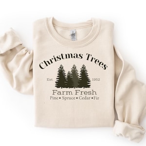 Könnte beinhalten: Ein cremefarbenes Sweatshirt mit Rundhalsausschnitt und einer dunkelgrünen Grafik von drei Tannenbäumen. Der Text lautet: Christmas Trees, Est. 1952, Farm Fresh, Pine, Spruce, Cedar, Fir. Das Design ist mittig auf der Brust des langärmeligen Oberteils.