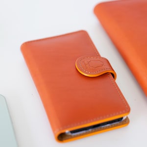 Op de afbeelding: Een rechthoekig oranje lederen telefoonhoesje met een veilige drukknoopsluiting. De flap is voorzien van een klein in reliëf gemaakt schildlogo met de tekst "gohuni". Het hoesje heeft zichtbare stiksels langs de randen en een gladde afwerking.