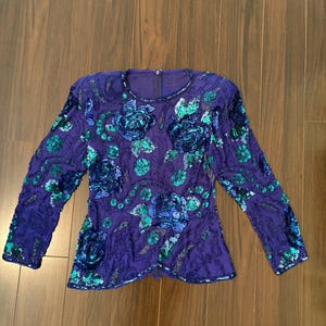 Op de afbeelding: Een top met lange mouwen in dieppaars, zwaar versierd met iriserende blauwe en groene lovertjes in bloemmotieven. Het kledingstuk heeft een ronde halslijn, een rechte zoom en een verticale ritssluiting in het midden van de rug.