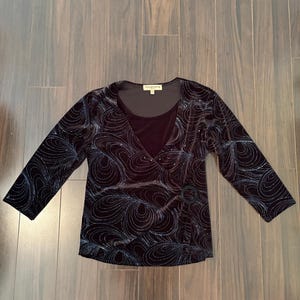 Könnte beinhalten: Ein schwarzes Langarmshirt mit V-Ausschnitt und einer Wickeloptik-Vorderseite, die an der Taille mit einem runden Ring verziert ist. Der Stoff zeigt ein dunkles, wirbelndes Muster in schimmernden Blau- und Silbertönen. Das Etikett zeigt Notations.