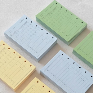 May include: A collection of rectangular paper planner inserts in pastel yellow, light blue, and sage green. Each sheet features a printed monthly calendar grid, a section for notes, and six pre-punched holes along the side for binder storage.