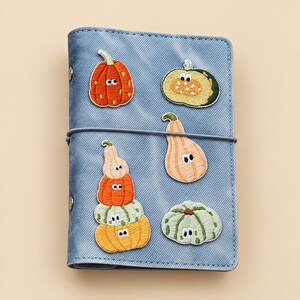 Könnte beinhalten: Ein hellblauer Notizbuchumschlag aus Stoff mit gestickten Aufnähern verschiedener Kürbisse und Zierkürbisse mit Cartoon-Augen. Der Umschlag wird mit einem dünnen Gummiband zusammengehalten und hat zwei sichtbare Metallnieten an der linken Seite.