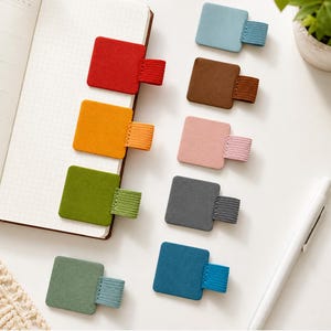 May include: A collection of nine square adhesive pen holders in various colors including red, orange, green, light green, blue, brown, pink, dark gray, and teal. Each holder features a matching elastic loop attached to a square base.