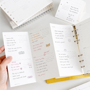 May include: A white multi-fold planner insert with black text and handwritten notes in various colors. The pages feature monthly calendars, checklists, and small illustrations like hearts, a sun, and a bear. The planner is held by a metal ring binder.