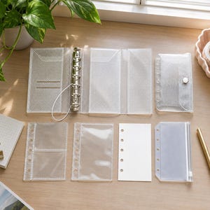 Puede incluir: Una funda de carpeta de seis anillas de plástico transparente texturizado con cierre elástico, acompañada de un estuche a juego con botón a presión y varios insertos transparentes, incluyendo un tarjetero, un bolsillo liso y una funda con cremallera.