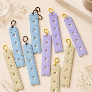 Puede incluir: Una colección de ocho reglas rectangulares de plástico en colores pastel azul, lavanda y crema. Cada regla presenta una escala métrica vertical, una serie de recortes circulares y una pequeña cadena de cuentas unida a la parte superior. El texto Long Lab está impreso en cada pieza.