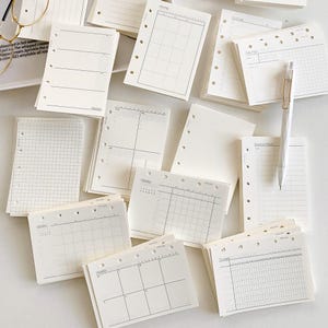 May include: A collection of off-white loose-leaf planner inserts with various layouts including monthly calendars, weekly schedules, grid paper, and trackers. Each sheet features pre-punched holes along the side for binding and a white ballpoint pen.