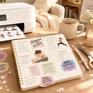 May include: A spiral-bound planner open on a desk, featuring handwritten workout plans, daily affirmations, and to-do lists. Decorative stickers include text like STRONGER THAN YESTERDAY and MAKE YOURSELF PROUD, alongside illustrations of a person doing yoga.