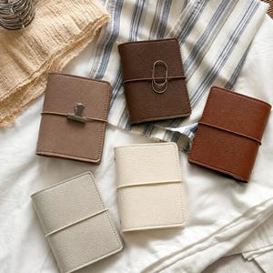 May include: Five small rectangular notebooks with textured covers in shades of beige, light brown, dark brown, and cream. Each notebook is secured with a thin elastic band wrapped horizontally around the center, featuring metal clips or paper clips.