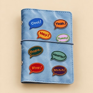 Peut inclure: Une couverture de carnet bleu clair à texture denim ornée de huit écussons brodés en forme de bulles de dialogue. Les écussons portent les mots Cool!, Yeah!, Hey!, Yummy!, Oops, Hooray!, Wow! et Sh-h-h! dans des fils de couleurs vives.