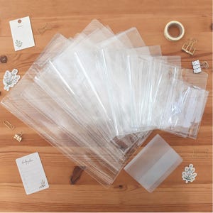 May include: A collection of clear plastic document sleeves and a translucent frosted folder arranged in a fan shape, accompanied by small gold-toned metal paper clips, a roll of adhesive tape, and two small rectangular cards with floral illustrations.