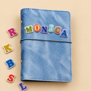 以下が含まれることがあります： デニム調の青いノートカバー。カラフルな刺繍の布製文字パッチで「MONICA」と綴られています。その横には、R、K、B、S、Lなど、他にもバラバラのカラフルな刺繍文字パッチがいくつか置かれています。