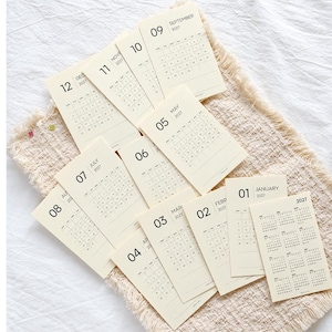 May include: A collection of twelve individual cream-colored paper calendar cards for the year 2027. Each card features a large month number, the month name, and a grid of dates. One card displays the full 2027 annual calendar overview.