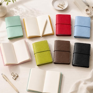 May include: A collection of small, rectangular notebooks in various colors including mint green, yellow, red, light blue, lime green, brown, black, and pink. Each notebook features a textured cover and an elastic band closure. Some are open to show blank or lined pages, accompanied by a wooden pencil and metal binder clips.