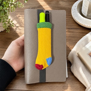 May include: A gray leather-textured notebook featuring a vertical dark gray elastic band. Attached to the band is a small, ribbed knit sock-shaped pen holder in yellow with a green cuff, a blue heel, and a red toe, containing a black and neon pen.