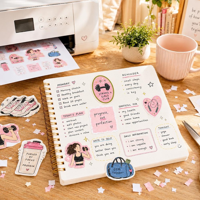 May include: A spiral-bound planner open to a page with handwritten daily tasks, affirmations, and fitness-themed stickers. The page includes sections for Monday, reminders, gratitude, and daily affirmations, decorated with pink and blue illustrations.