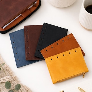 May include: A collection of five rectangular leather pouches in blue, dark brown, black, tan, and yellow. Each pouch features a row of punched holes along one edge for binding, and the yellow pouch includes a metal zipper closure at the bottom.
