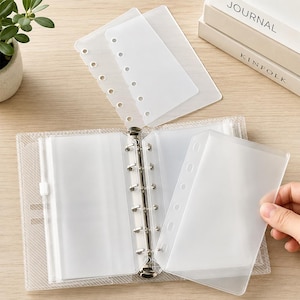 May include: A clear, six-ring binder with a textured cover and multiple transparent plastic insert pages. The binder features a metal ring mechanism for organizing loose sheets, accompanied by several loose, translucent, pre-punched plastic dividers.