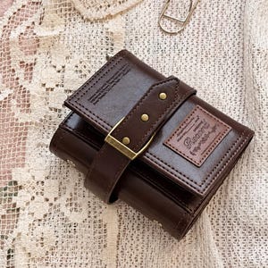May include: A dark brown wallet featuring a decorative strap with a brass-toned buckle and studs. The front flap displays a rectangular patch with the embossed text "Record" and additional small printed text details on the upper left corner.
