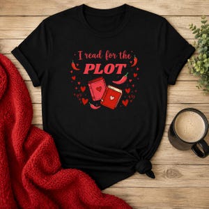May include: A black short-sleeved t-shirt featuring a graphic print with the text I read for the PLOT in red letters. The design includes two red books with heart icons, several small red hearts, and red chili pepper illustrations scattered around.