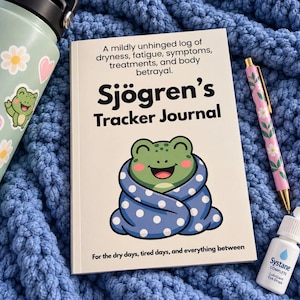 May include: A white journal titled Sjögren's Tracker Journal featuring a green frog wrapped in a blue polka dot blanket. The cover text reads: A mildly unhinged log of dryness, fatigue, symptoms, treatments, and body betrayal. For the dry days, tired days, and everything between.