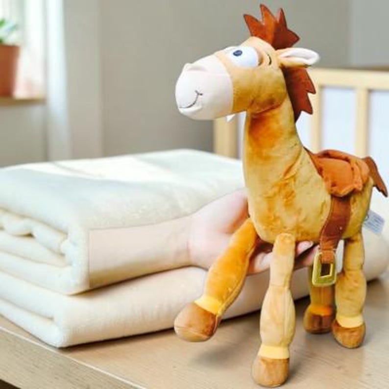 May include: A plush toy horse with a tan body, a dark brown mane, and a brown saddle featuring a gold-colored buckle. The horse has large white eyes with black pupils, a light-colored muzzle, and a stitched smile. The toy is held near a folded cream-colored blanket.