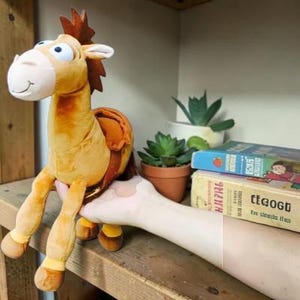May include: A plush horse toy with a tan body, a dark brown mane, and a light-colored muzzle. The stuffed animal features large white eyes with black pupils, a stitched smile, and a brown saddle strapped around its midsection.