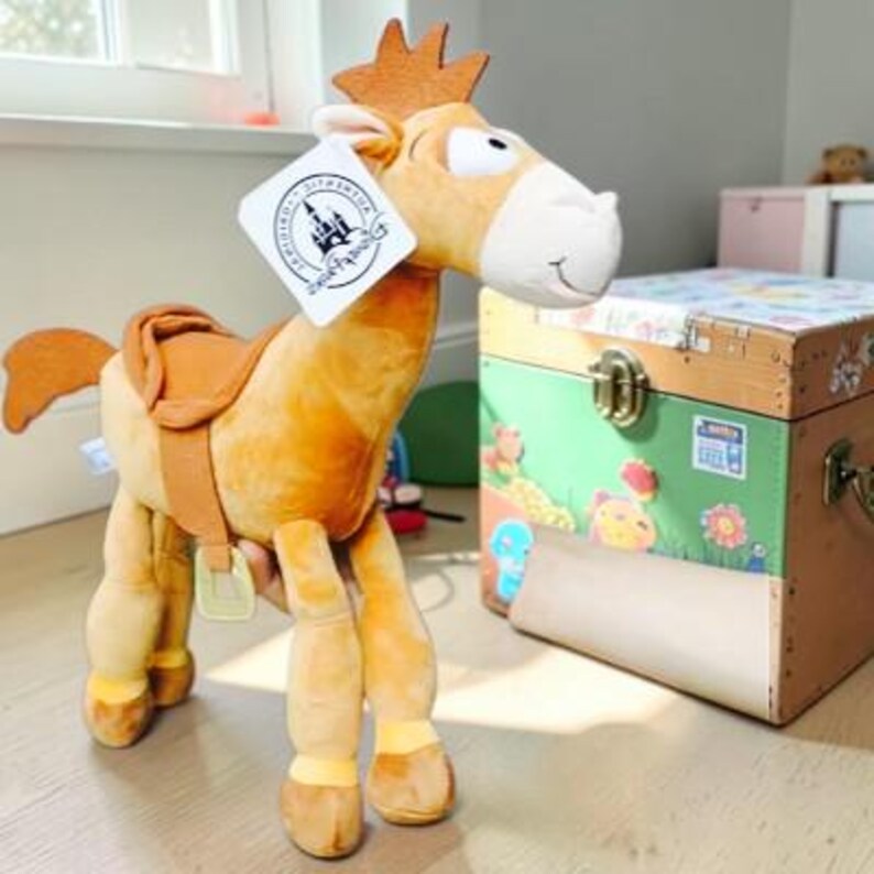 May include: A tan plush horse toy with a white muzzle, a brown mane, and a brown saddle. The toy features a white rectangular tag attached to the ear with a black Disney logo and the text Authentic Original. The horse has four legs and a tail.