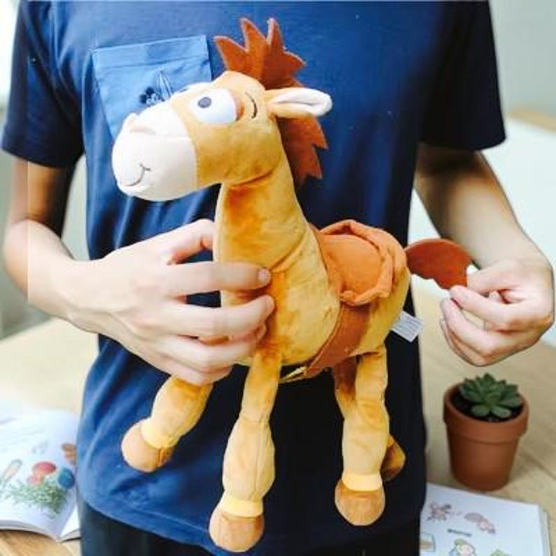 May include: A plush horse toy featuring a tan body, a dark brown mane, and a matching brown saddle. The stuffed animal has large white eyes with black pupils, a light-colored muzzle, and four legs with darker brown hooves at the bottom ends.