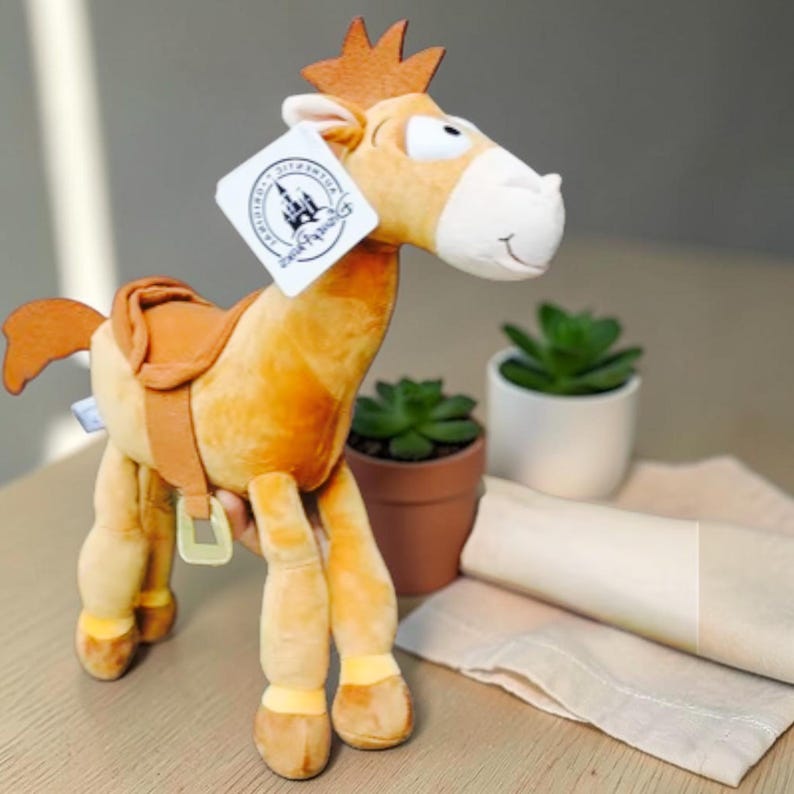 May include: A plush toy of a tan horse with a brown mane, tail, and saddle. The horse has a white muzzle and a white tag attached to its ear featuring a castle logo and the text Authentic Disney Parks. The toy has four legs and a soft texture.