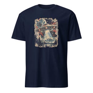 May include: A navy blue short-sleeve t-shirt featuring a graphic print of a flying saucer hovering over a mountain landscape with pine trees and a large moon. The design uses a distressed, textured aesthetic with muted red, blue, and cream tones.