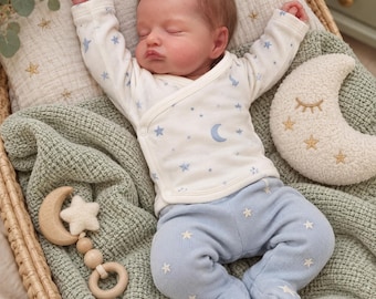 Könnte beinhalten: Eine realistische Babypuppe mit braunen Haaren schläft in einem Weidenkorb. Die Puppe trägt ein weißes Langarm-Wickelshirt mit blauen Monden und Sternen sowie eine hellblaue Strampelhose mit weißer Sternenstickerei. Ein Mondkissen und eine Holzrassel liegen bei.