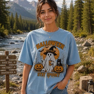 Peut inclure: Un t-shirt bleu clair avec un motif représentant un fantôme portant un chapeau de sorcière, tenant une boisson et faisant un signe de paix, entouré de deux citrouilles d'Halloween et d'un chat noir, avec le texte "HALLOWEEN" et "STAY SPOOKY".