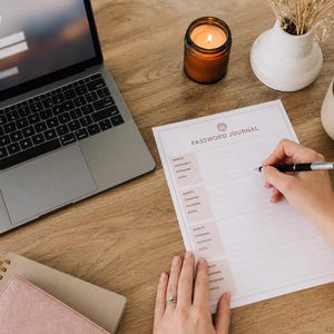 Könnte beinhalten: Ein ausgedrucktes Password Journal-Blatt mit hellrosa Abschnitten für Website, Benutzername, Passwort und Notizen, platziert neben einem Laptop, einer brennenden Kerze in einem braunen Glasgefäß, einer weißen Keramiktasse und einem rosafarbenen Spiralnotizbuch.