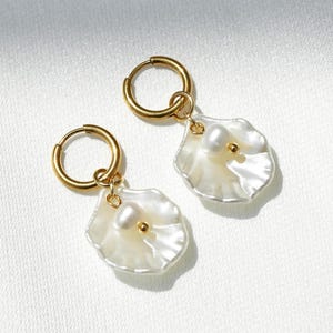 Op de afbeelding: Een paar gouden oorringen met witte, bloemblad-vormige hangers. Elke hanger heeft een kleine parel en een gouden accent. De oorbellen worden gepresenteerd op een witte, getextureerde ondergrond, wat een minimalistische uitstraling creëert.