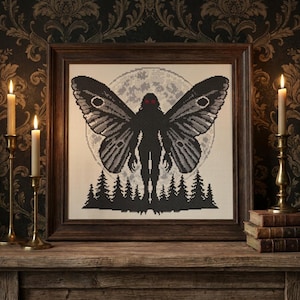 May include: A framed cross-stitch embroidery featuring a dark silhouette of a winged creature with glowing red eyes, positioned in front of a full moon and a forest of pine trees. The artwork is displayed in a dark wooden frame.