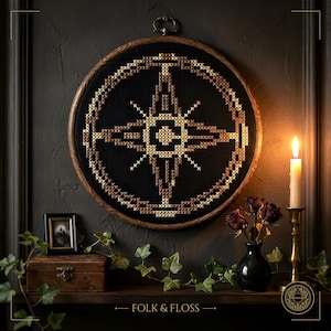 Può includere: Un telaio da ricamo circolare in legno contenente un disegno a punto croce di una rosa dei venti dorata su uno sfondo di tessuto nero. Il motivo presenta complessi schemi geometrici in tonalità oro e bronzo, centrati in un bordo circolare.