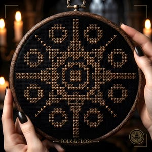 Può includere: Un telaio da ricamo circolare in legno contenente tessuto nero con un motivo a punto croce simmetrico in filo color bronzo metallizzato. Il design presenta un quadrato centrale circondato da forme geometriche. Il testo FOLK & FLOSS è visibile.