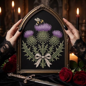 May include: A gothic-style arched frame containing a detailed cross-stitch embroidery on black fabric. The design features three purple thistle flowers with green leaves, a bumblebee resting on the center bloom, and a beige ribbon bow at the base.