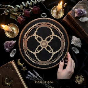 May include: A circular wooden embroidery hoop containing a cross-stitch pattern on black fabric. The design features a geometric, interlocking circular motif in shades of gold, bronze, and brown thread, centered within the dark fabric frame.