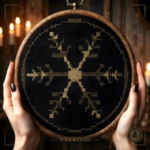 Può includere: Un telaio da ricamo circolare in legno contenente tessuto nero con un motivo a fiocco di neve a punto croce dorato. Il design presenta un quadrato centrale con rami radianti. Il testo FOLK & FLOSS è stampato nella parte inferiore.