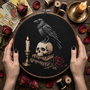 May include: A circular wooden embroidery hoop containing black fabric with a cross-stitch design of a grey raven perched on a human skull, which rests atop two stacked books next to a lit candle and a red rose, all rendered in detailed thread.